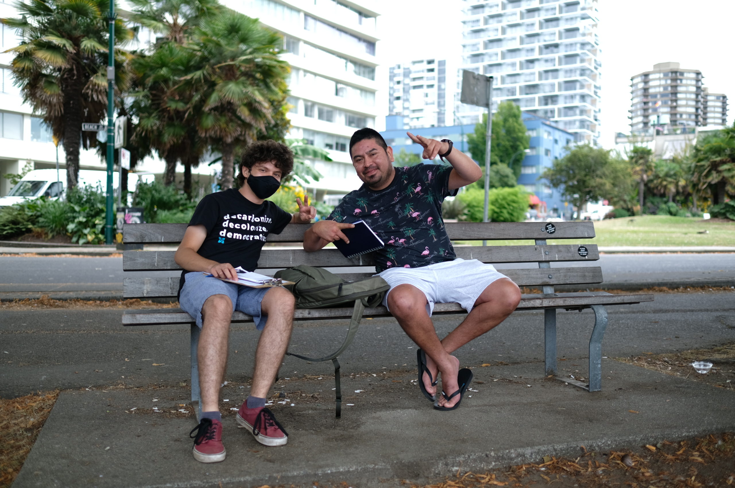 Two people sitting on a bunch smiling for the camera and doing sideways peace signs with their hands, buildings/city in the background.